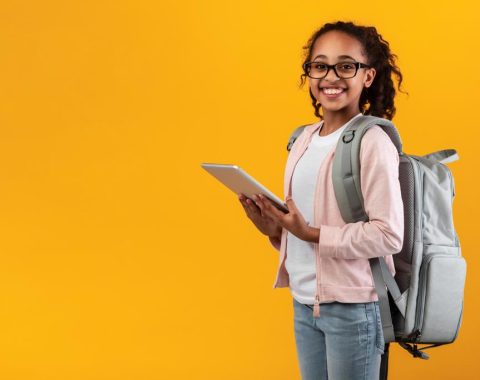 Online Classes Concept. Smiling teenage pupil wearing glasses and backpack holding and using digital tablet for studying standing on yellow orange studio wall posing at camera, free copy space