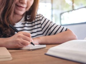 Closeup image of a woman writing on blank notebook with books on wooden table while learning