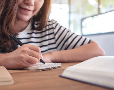 Closeup image of a woman writing on blank notebook with books on wooden table while learning