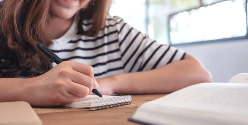 Closeup image of a woman writing on blank notebook with books on